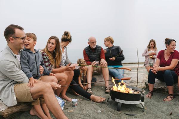 Eine Gruppe Menschen unterschiedlichen Alters sitzt am Strand im Halbkreis um ein Lagerfeuer herum.
