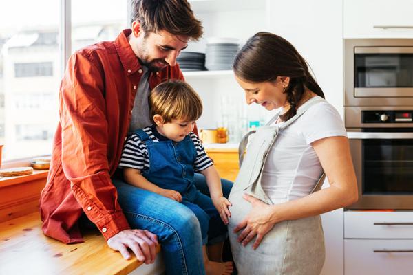 Ein Mann und ein kleiner Junge sitzen auf der Arbeitsplatte in der Küche und betrachten den Bauch einer schwangeren Frau, die vor ihnen steht.