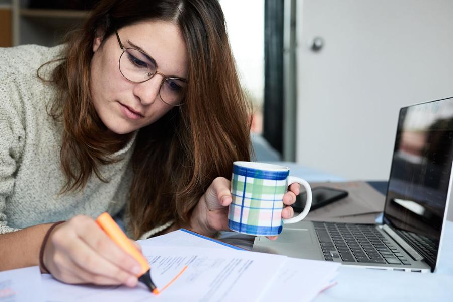 Eine Frau sitzt mit einer Tasse in der Hand vor einem Laptop und markiert auf einem Schriftstück eine Textpassage.