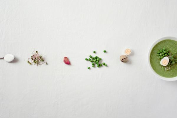 Nebeneinander aufgereiht von links nach rechts: ein Teelöffel mit Salz, Sprossen, eine Knoblauchzehe, grüne Erbsen, ein Wachtelei und eine Schale Erbsensuppe.