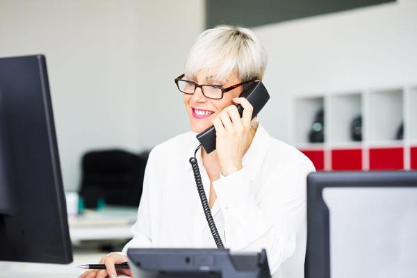 Eine lächelnde Frau mit kurzem Haar und Brille sitzt vor einem Computer und hält sich einen Telefonhörer ans Ohr.