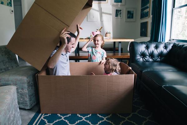 Ein Mann und ein Kleinkind sitzen in einem großen Pappkarton, daneben steht ein kleines Mädchen, alle drei lachen.